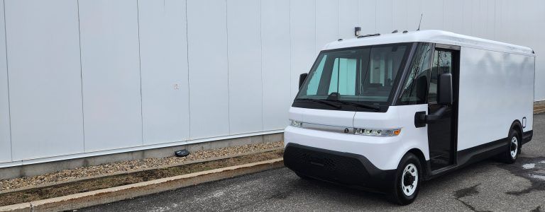 Une camionnette de livraison blanche est garée sur une surface pavée devant un grand mur uni. La camionnette est équipée de fonctionnalités de conception modernes.